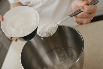 A close photo of the hands of a young woman who is tossing a spoonful of wheat flour into the stainless steel bowl of the mixer.