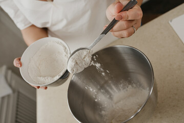A close photo of the falling wheat flour from the spoon in the hands of the young woman into the stainless steel bowl of the mixer.