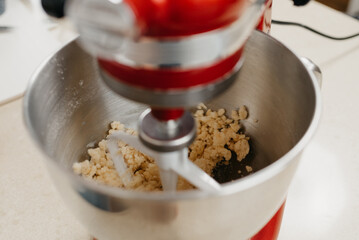 A close photo of a red stand mixer which is whipping butter, almond flour, wheat flour, powdered sugar, and yolk to the shortcrust pastry in a stainless steel mixing bowl in a kitchen.