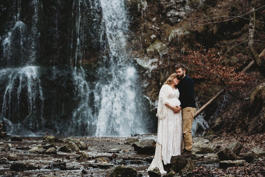 Husband and expecting wife outside in winter by a waterfall in winter - Powered by Adobe