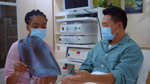 Asian shocked patient lying in bed communicating with nurse afroamerican doctor being confused about diagnosis checking lung xray image on doctor appointment.