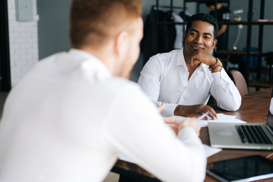 Pensive serious professional African American man interviewing caucasian European young candidate for job at successful company. Two business people talking strategy sitting at office desk.