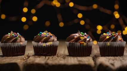 Tasty chocolate cupcakes with sprinkling on rustic wooden table on black background with yellow lights. Sweet dessert. Bakery concept. Elegant food. Sweets for coffee or tea. Space for text.