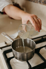 Fototapeta na wymiar A hand of a woman who is pouring fresh lemon juice from the glass to the saucepan on the gas stove