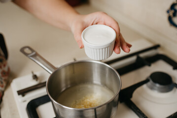 A close photo of a hand of a woman who is preparing to pour cream from the cup to the saucepan with lemon juice and zest on the gas stove