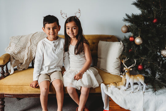 Multiracial young girl & boy sitting on couch next to christmas decor