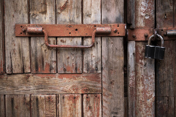 Padlock and rusty metal handles on the gate made of old, textured wood with peeling paint.The...