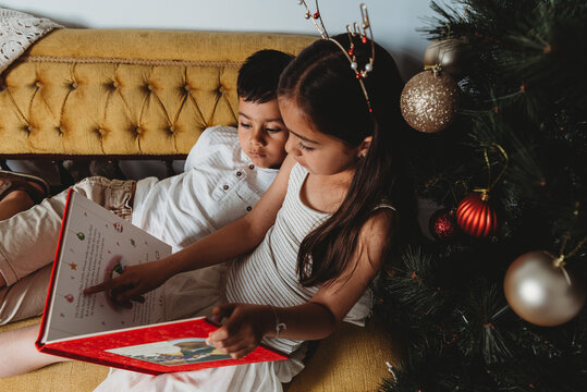 Multiracial young girl & boy reading christmas book on lounge