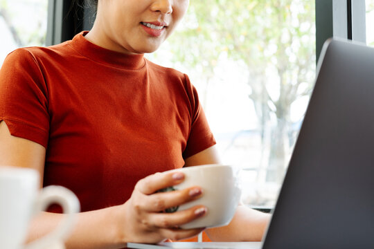 Asia woman hold cup of coffee while typing on laptop keyboard. W