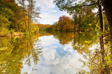 The autumn-colored forest frames the lake, in which the clouds are reflected.