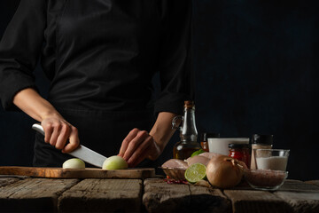 Close-up view of chef cuts with knife onion on wooden chopped board. Backstage of cooking traditional Indian chicken curry on dark blue background. Frozen motion. Concept of cooking tasty hot meal.