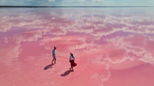 cirle drone flight over lovely couple tourists with corgi dog walking on bright beautiful landscape with beautiful pink lake and clouds reflections on water surface