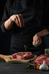 Close-up view of chef in black uniform pours rosemary on raw steak on wooden chopped board. Backstage of preparing grilled pork meat at restaurant kitchen on dark blue background. Frozen motion.