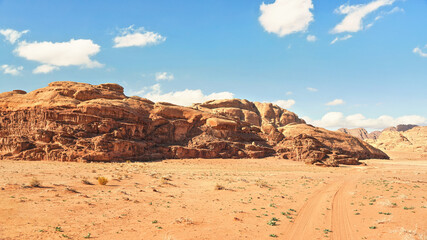 Fototapeta na wymiar Rocky massifs on red sand desert, vehicle tracks ground, bright cloudy sky in background, typical scenery in Wadi Rum, Jordan