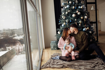Obraz na płótnie Canvas Lovely family sit near the Christmas tree at home, caring mom hold little daughter in arms, loving husband kiss beautiful wife, enjoy winter holidays, New Year celebration concept