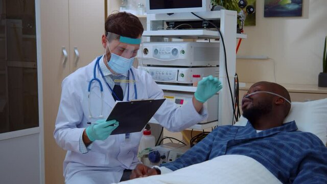 Caucasian man in face shield doctor consulting young poor african american sick man suffering coronavirus infection in hospital quarantine concept.