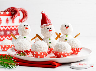 Snowmen made of coconut balls in santa hat. Healthy christmas dessert. Funny and cute new year sweets. Festive food. Winter and christmas concept. Fir branches, candy cane.White background. Copy space