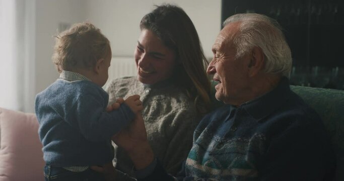 Cinematic shot of happy family: grandfather, daughter and grandson baby are having fun to pass time together while sitting on sofa at home. Concept of life, grandparents, generation, childhood, love