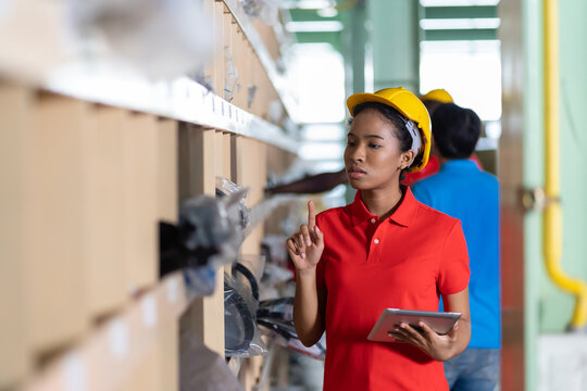 African American Female Warehouse Worker In Red Uniform And Helmet Working And Checking Automotive Spare Parts On Shelf Pallet In Storage Automotive Spare Parts Warehouse