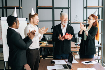 Successful business team of multi-ethnic people giving present to happy mature adult male boss. Multinational businessteam wearing festive holiday hats applauding to mature adult male leader at office