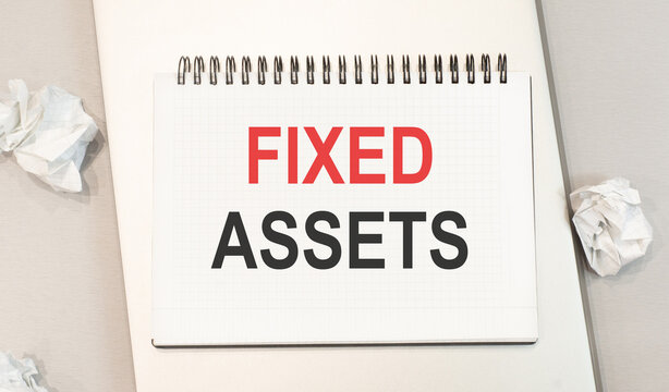 The word fixed assets is written in a notebook that sits on a gray desktop along with a laptop. Business image