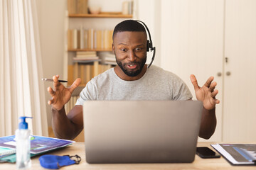 African american man using phone headset while having a video chat on laptop while working from home