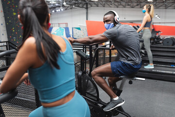 Fit african american man and fit caucasian woman wearing face masks exercising on stationary bike in
