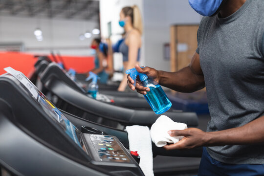 Mid section of fit african american man wearing face mask sanitizing cardio machine before working o