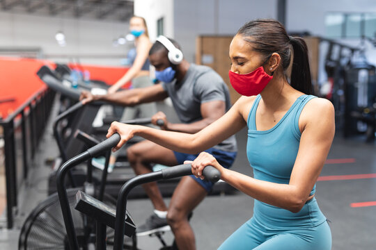 Fit african american man and fit caucasian woman wearing face masks exercising on stationary bike in