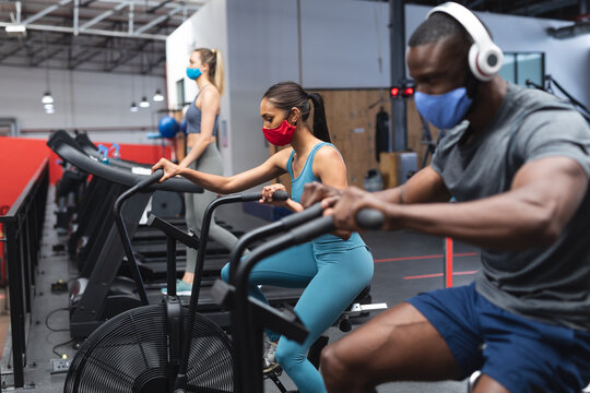 Fit african american man and fit caucasian woman wearing face masks exercising on stationary bike in