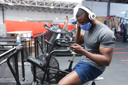 Fit african american man wearing face mask and headphones using smartphone while sitting on stationa