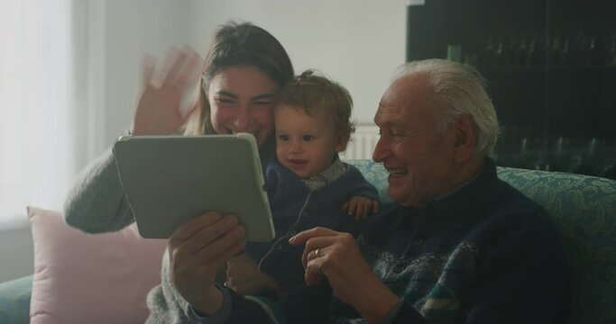 Cinematic shot of happy family: grandfather, daughter and grandson baby having fun to make selfie or video technology call to family and relatives with tablet or smartphone on sofa in living room at h