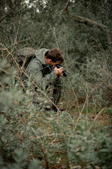 young mountaineer man dressed in dark green in the undergrowth crouching taking photos of the plants, dark and desaturated style, adventure concept.