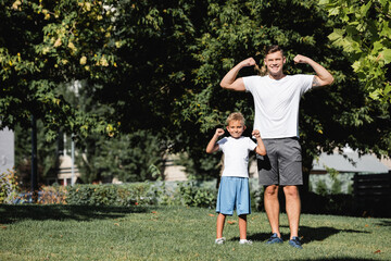 Fototapeta premium Smiling father and son looking at camera while posing with raised hands showing power in park