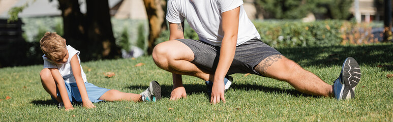 Preschooler boy in sportswear doing lunges near young adult man in park on blurred background, banner