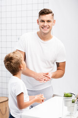 Obraz premium Son holding soap and standing near smiling father rubbing hands while looking at camera in bathroom
