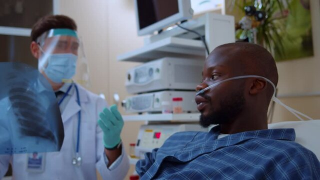 Coronavirus patient afro-american sick man lying on hospital bed talking to caucasian doctor wearing face mask shiled social distance discussing disease. Pandemic. Quarantine.