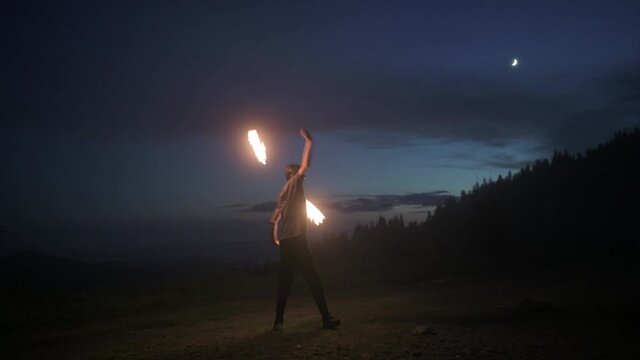 Graceful male fireshow performer making fire dance with two metal fans lit with flames on night mountains meadow
