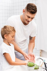 Obraz na płótnie Canvas Smiling father and son washing hands, while standing near sink in bathroom