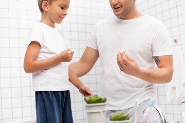 Smiling son standing near father and looking at dental floss holder in bathroom on blurred foreground