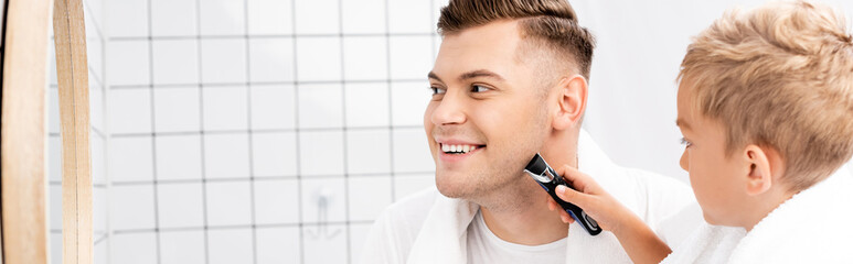 Son holding electric razor and shaving face of father smiling and looking at mirror, banner