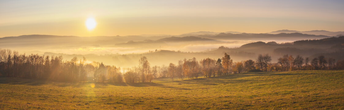 sunset in the mountains - hilly landscape with meadows and forests in a haze, into a yellow and orange-colored sky © Roman
