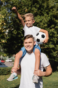 Happy son with hand in air holding ball, while riding piggyback on smiling father in park on blurred background
