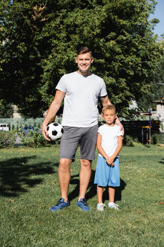 Smiling man in sportswear with ball embracing preschooler boy with clenched hands in park