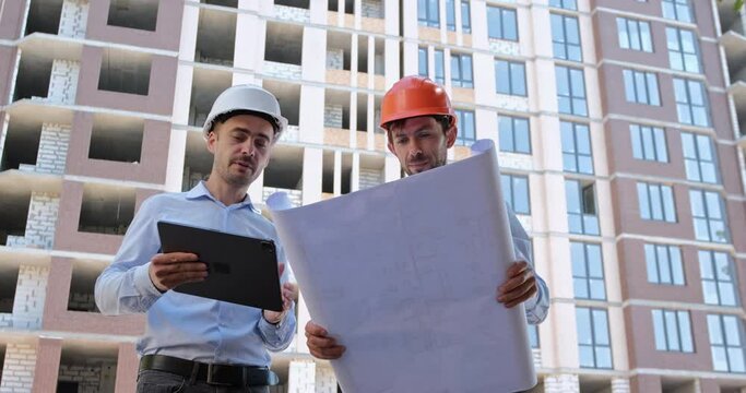 An engineer with a tablet computer convinces the builder with a paper drawing of the construction correctness. Builders actively communicate against the backdrop of a house under construction.