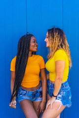 Lifestyle, teenage girl friends reflecting love with each other on a blue wall background dressed in yellow t-shirts. Black girl with long braids and blonde Caucasian girl.