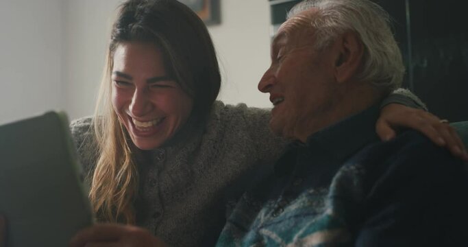 Cinematic shot of happy granddaughter and grandfather are having fun to make a selfie or video technology call to family and relatives with tablet while sitting on sofa in living room at home.