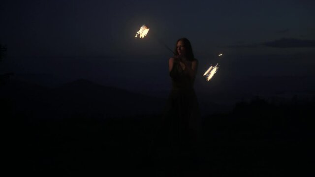 Graceful elegant red haired female fireshow performer making fire dance with two metal fans lit with flames on night mountains meadow