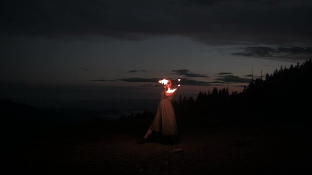 Graceful elegant red haired female fireshow performer making fire dance with two metal fans lit with flames on night mountains meadow