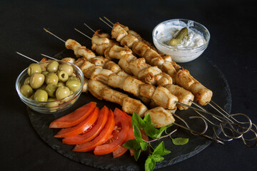 skewer of squid fillet on metal skewers on a dark background with sauce, olives and sliced tomatoes